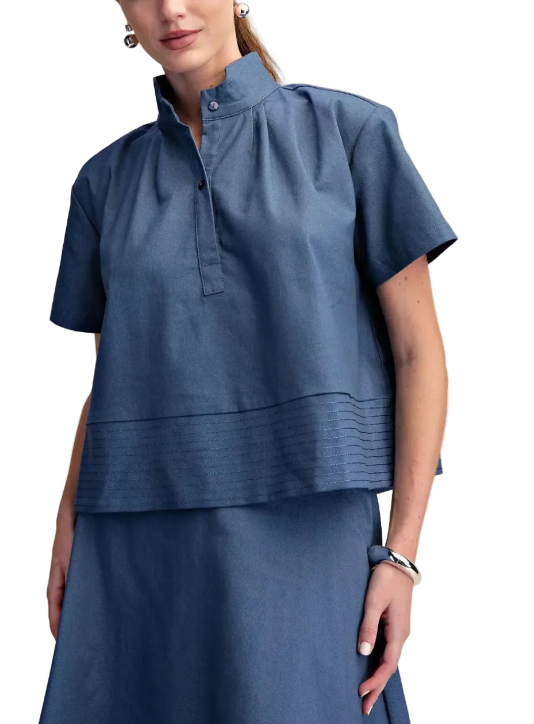 The front of the blouse is adorned with delicate pleats that cascade from the collar, creating a subtle yet eye-catching detail. A functional button-down placket allows for adjustable styling and comfort. The hem of the top is accentuated by a stylish band with intricate stitching, adding a refined finish and a hint of texture. This piece effortlessly blends classic tailoring with modern flair, making it perfect for both casual outings and more dressed-up occasions.

