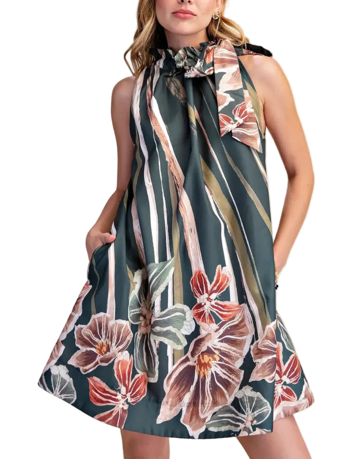 A woman modeling a sleeveless halter-neck A-line dress with a dark teal background and a large floral print in pink, brown, and white, set against a solid beige studio background.