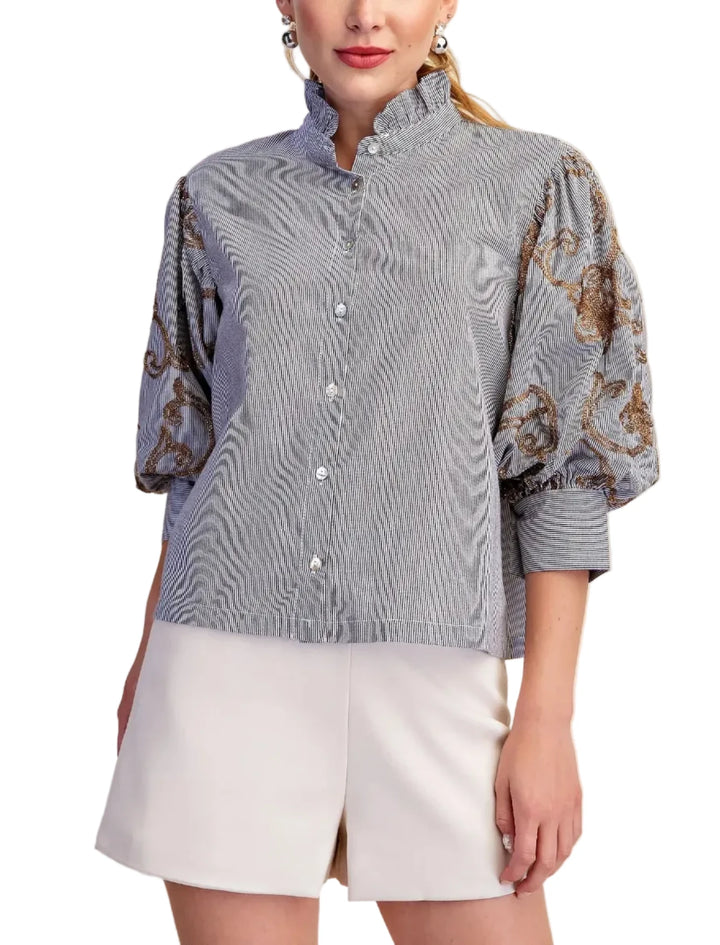 A woman modeling a light blue-gray chambray button-up blouse with a high stand collar, 3/4 puff sleeves, and brown paisley embroidery on the sleeves, paired with white shorts against a beige background.