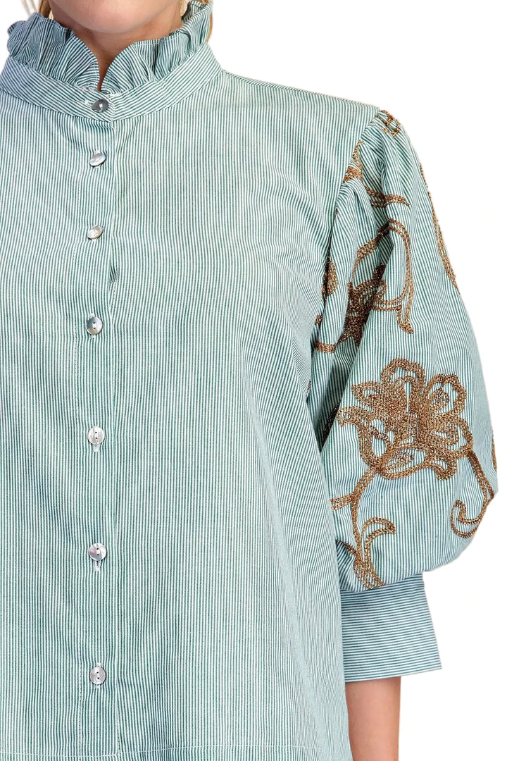 A close-up of a light blue and white striped blouse with a ruffled mock neck, button front, and voluminous sleeves featuring brown floral embroidery.