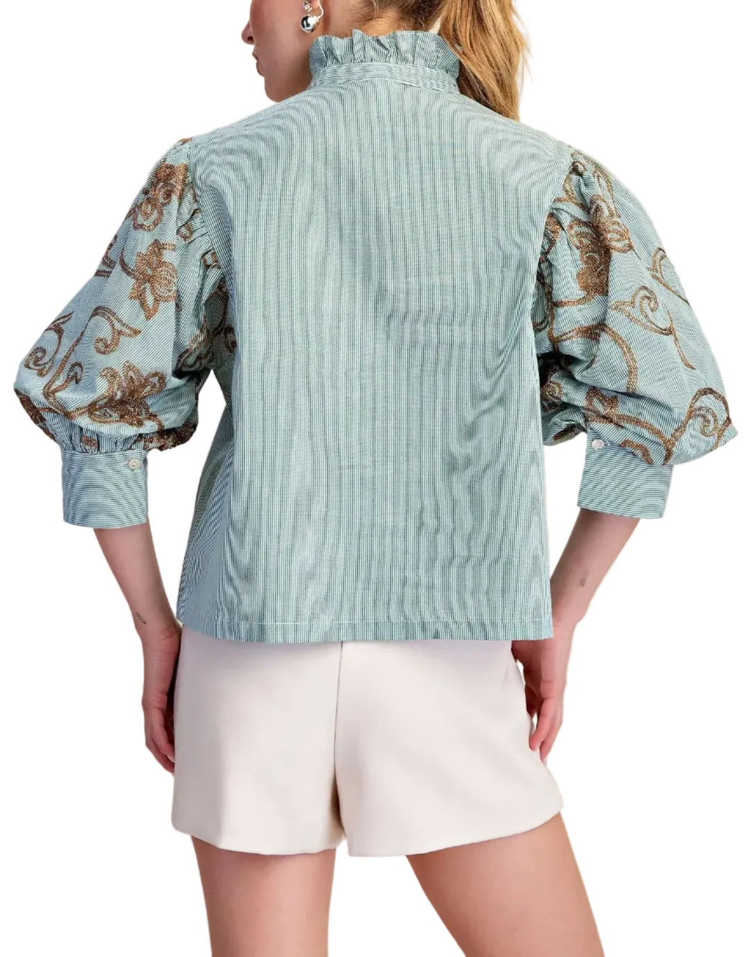 A woman shown from the back wearing a light blue blouse with ornate brown and gold patterned puff sleeves and white shorts, against a plain beige studio background.