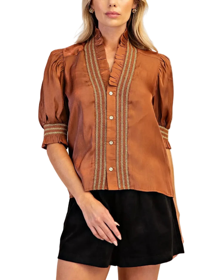 A woman modeling a brown short-sleeve blouse with vertical striped button placket and cuffs, paired with black shorts, against a plain beige studio background.