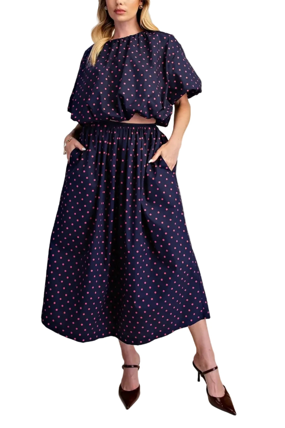 A woman modeling a navy blue two-piece set with a white and pink polka dot pattern, featuring a short-sleeve top with puff sleeves and a matching high-waisted skirt with side pockets, paired with dark brown pointed-toe heels against a solid beige background.