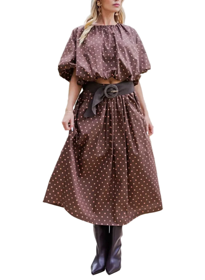 A woman stands outdoors on a paved walkway wearing a brown midi dress with white polka dots, a dark wide belt with a large buckle, and dark brown pointed boots.