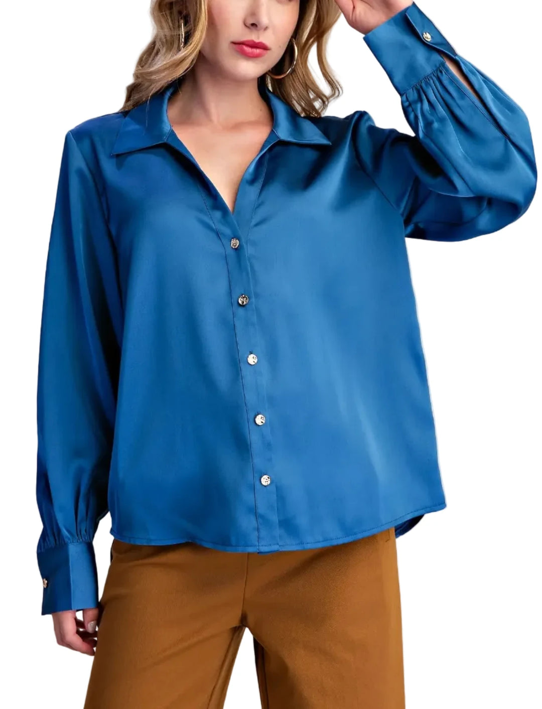 A woman wearing a long-sleeve satin-like button-up blouse in a rich medium blue, paired with brown pants, against a plain light beige studio background.