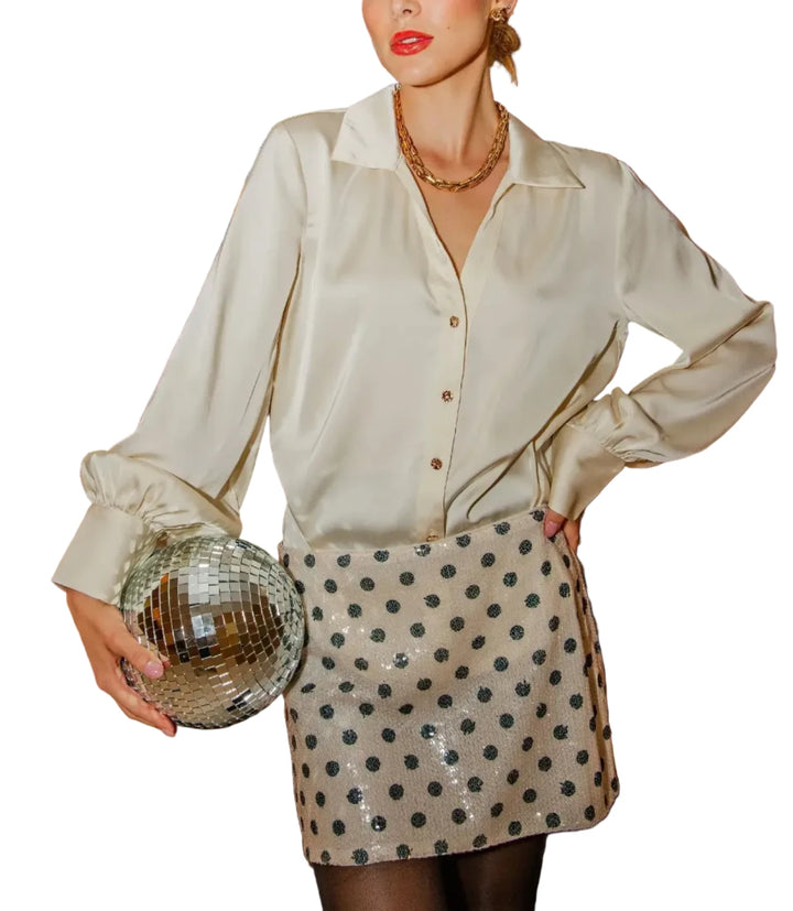 A woman models a cream satin button-up shirt and a cream sequined mini skirt with black polka dots, holding a silver disco ball clutch in front of a gold clothing rack.