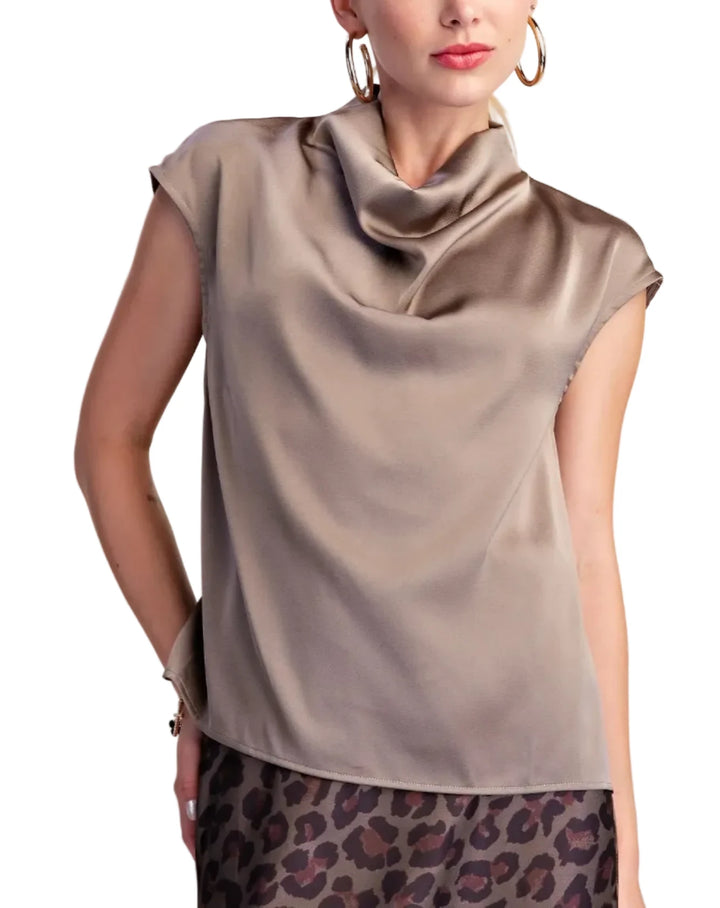A woman wearing a taupe satin-like cowl-neck top with cap sleeves and a leopard-print skirt, photographed against a plain light beige studio background.