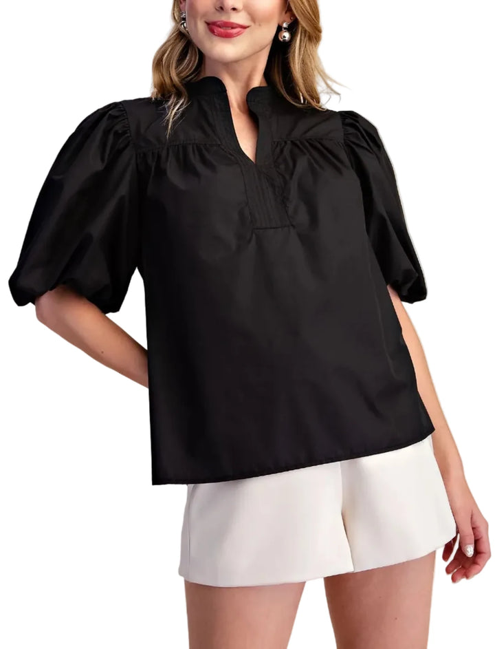 A woman wearing a black short puff-sleeve blouse with a mock neck and white shorts, standing against a plain beige studio background.