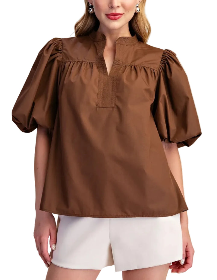 A woman modeling a brown short-sleeve blouse with puff sleeves, a gathered mock neck, and a small chest pocket, paired with white shorts against a plain beige studio background.