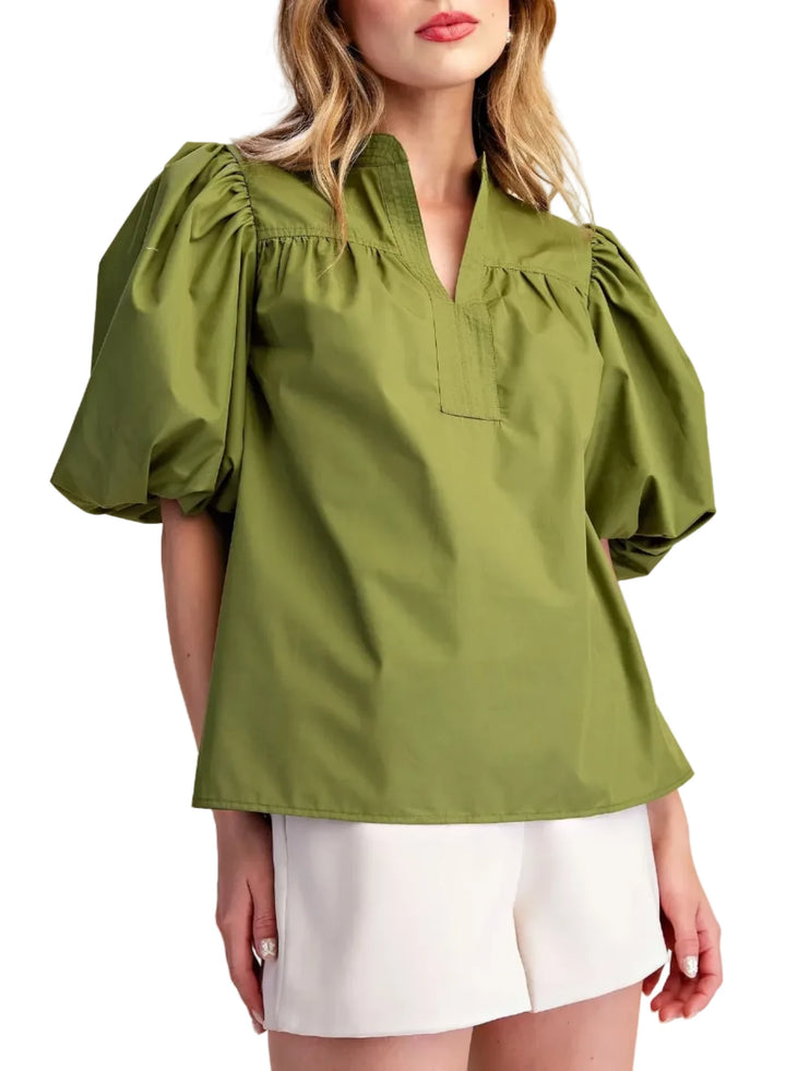 A woman wearing an olive green puff-sleeve blouse with a split neckline and white shorts, standing against a plain beige studio background.