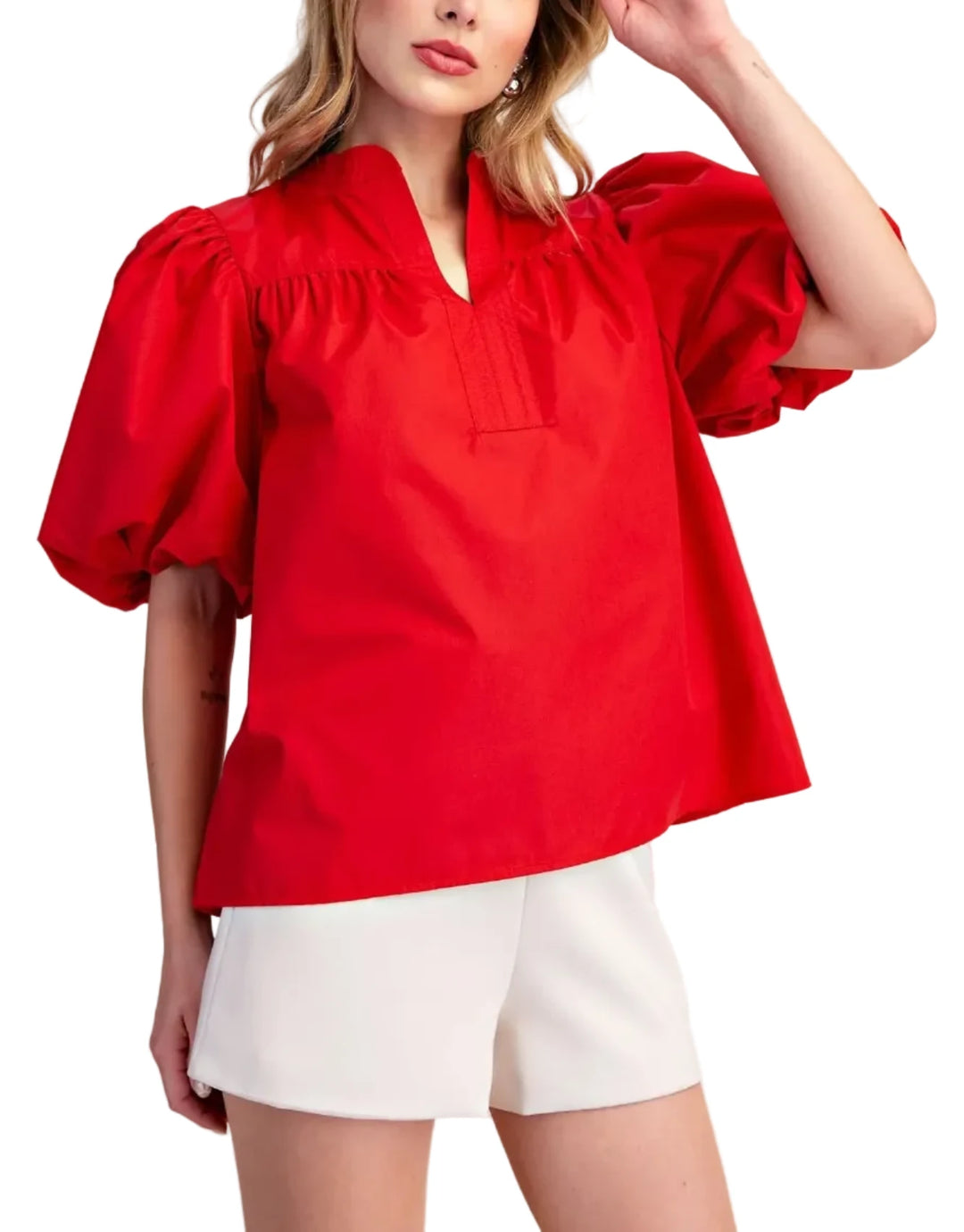 A woman modeling a bright red short-sleeve blouse with voluminous puff sleeves and a split neckline, paired with white shorts, against a plain beige studio background.