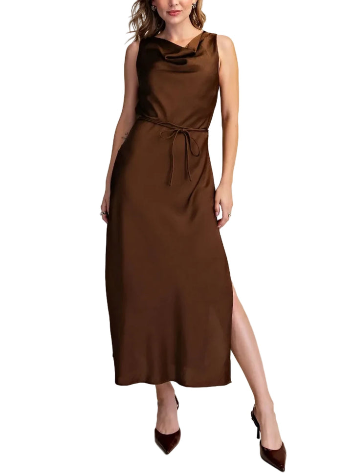 A woman models a sleeveless cowl neck midi dress in a rich chocolate brown satin fabric, featuring a tie waist and a high side slit, set against a solid beige studio background.