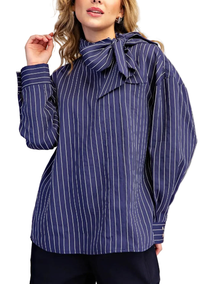 A woman wearing a navy blue long-sleeve blouse with thin white vertical pinstripes and a large bow detail at the high neckline, paired with dark pants, against a plain beige studio background.