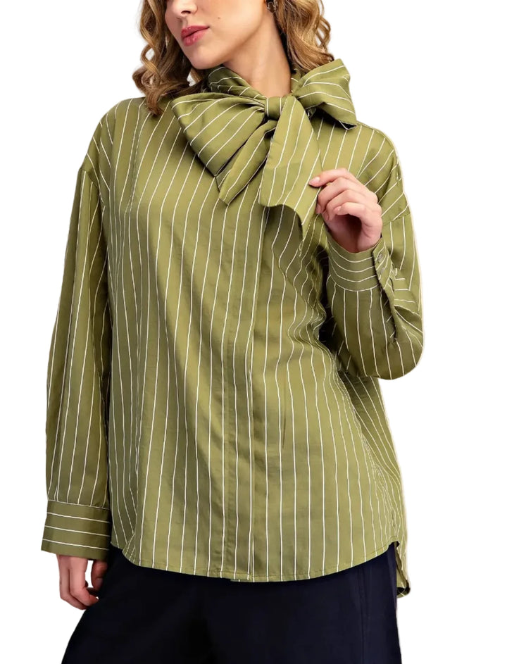 A woman wearing a sage green long-sleeve blouse with thin white vertical stripes and a large bow detail at the collar, paired with dark pants, set against a plain beige studio background.