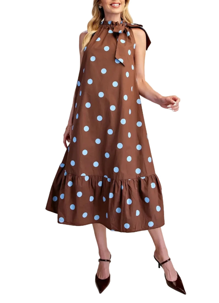 A woman models a sleeveless brown midi dress with light blue polka dots, a high ruffled collar with tie details, and a tiered hem, paired with dark brown pointed-toe heels against a solid beige studio background.