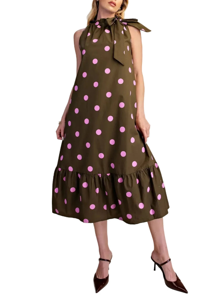 A woman modeling a sleeveless olive green midi dress with pink polka dots, a tie-neck bow, and a ruffled hem, paired with dark brown pointed-toe heels against a plain beige studio background.