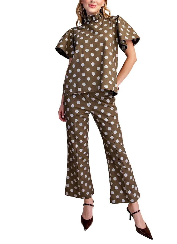 A woman models a matching two-piece set featuring a brown base with white polka dots, consisting of a short-sleeve mock-neck top with ruffled shoulders and matching wide-leg cropped pants, styled with dark brown pointed-toe heels against a solid beige studio background.