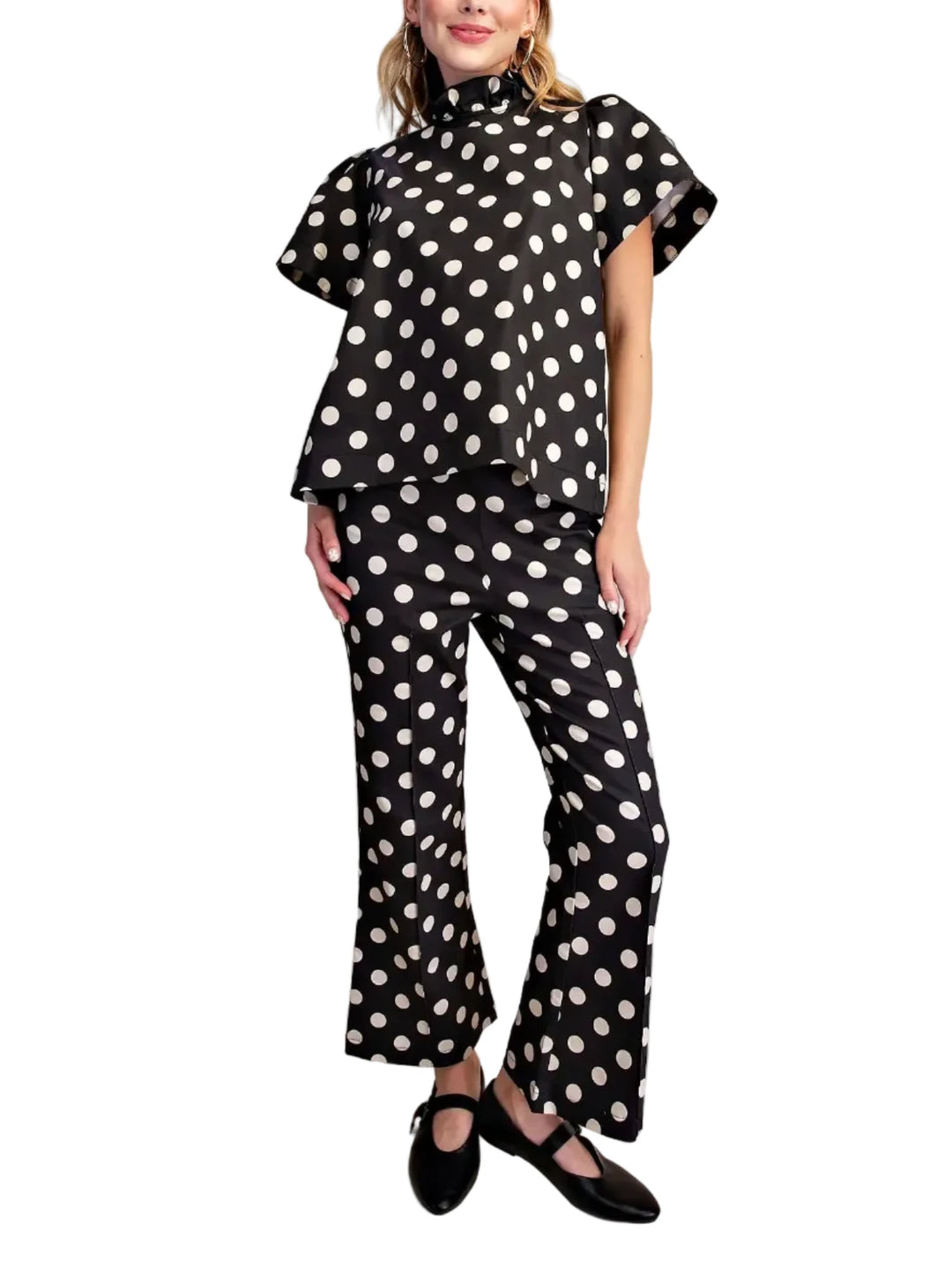 A woman modeling a black two-piece set with white polka dots, featuring a short-sleeve mock-neck top and matching flared pants, paired with black Mary Jane shoes against a plain beige studio background.