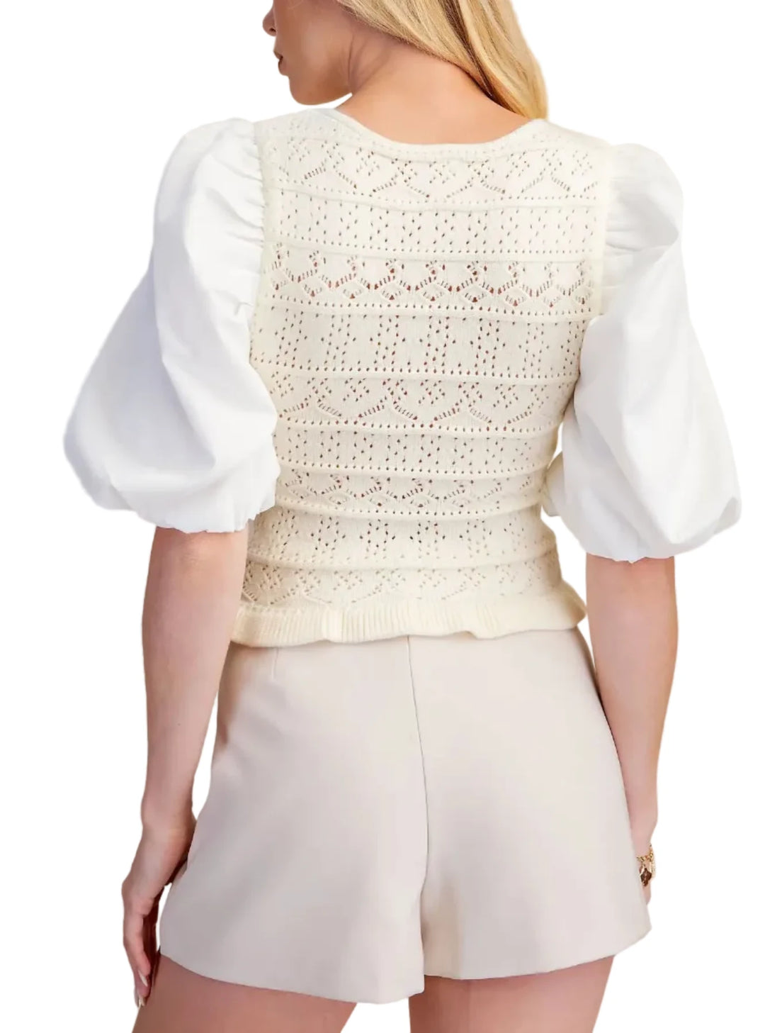A woman shown from the back wearing a cream-colored crochet knit vest with dramatic puff sleeves and cream shorts, set against a solid beige background.