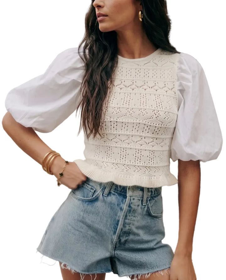 A woman wearing a white knit top with dramatic puff sleeves and light blue denim shorts, standing against a beige stone wall.