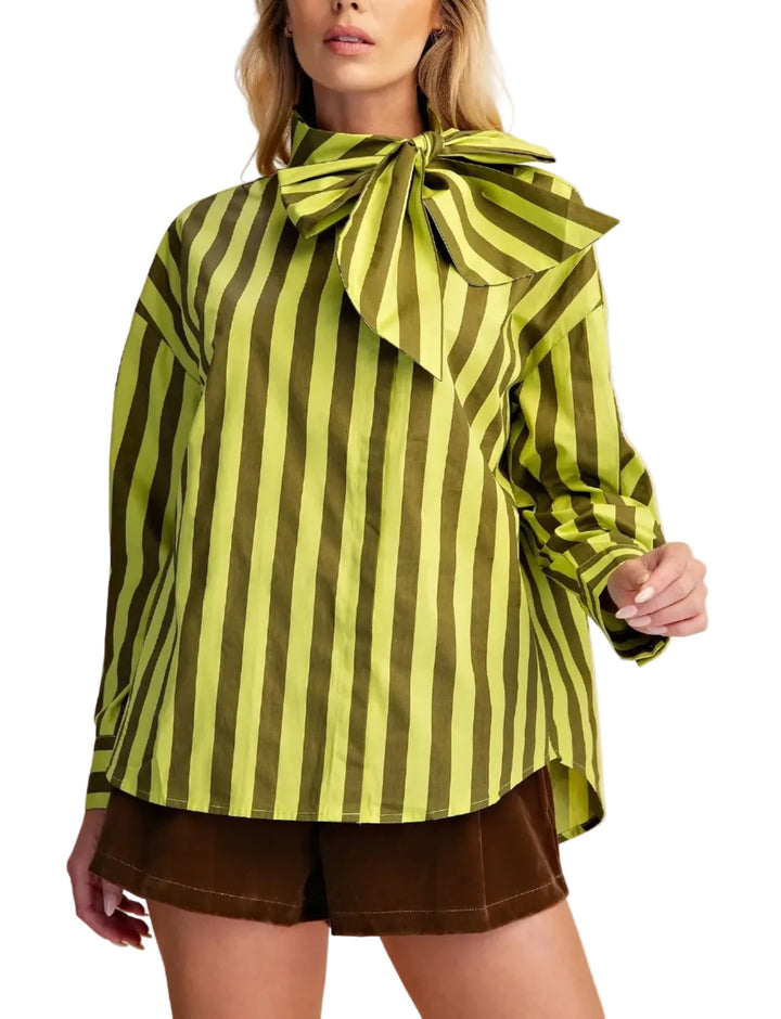 A woman modeling a lime green and olive green vertically striped blouse with a large bow at the collar, paired with brown shorts, against a solid beige background.