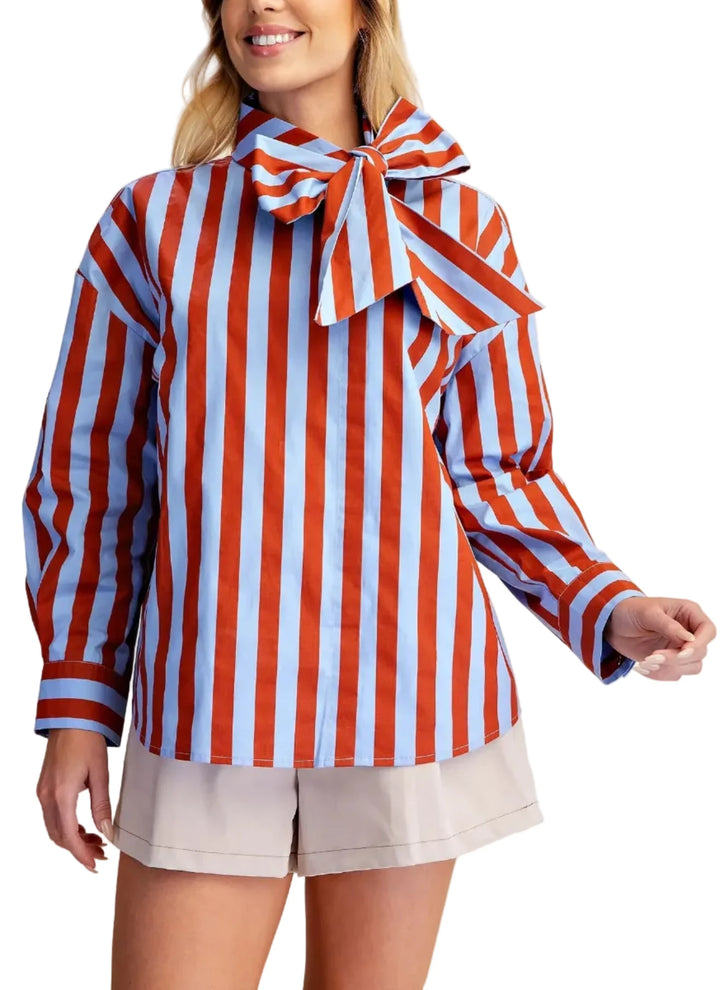 A woman wearing a long-sleeve striped blouse with red, white, and light blue vertical stripes, featuring a large bow at the collar, paired with light beige shorts against a solid beige background.