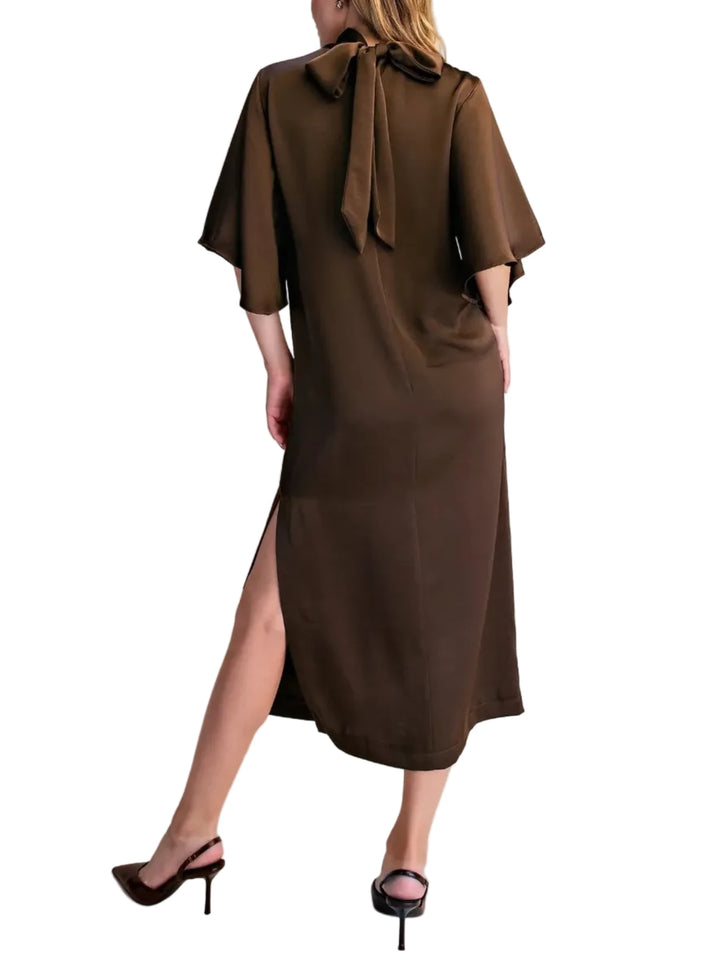 A woman modeling a brown midi dress from the back, featuring a large bow at the collar, short wide sleeves, and a side slit, paired with black heels against a plain beige background.