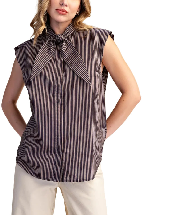 A woman modeling a sleeveless button-front blouse with a large bow tie neckline, featuring vertical dark brown and beige stripes, paired with light beige pants against a solid beige background.