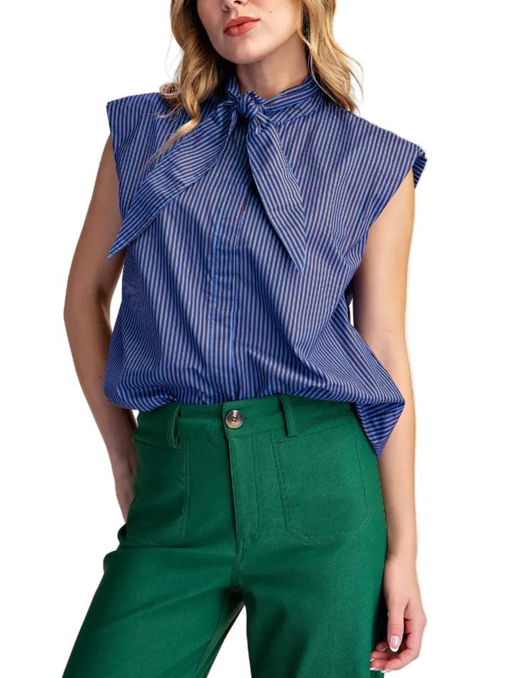 A woman wearing a sleeveless blue and white striped blouse with a large front bow tie collar, paired with emerald green pants, against a solid beige background.