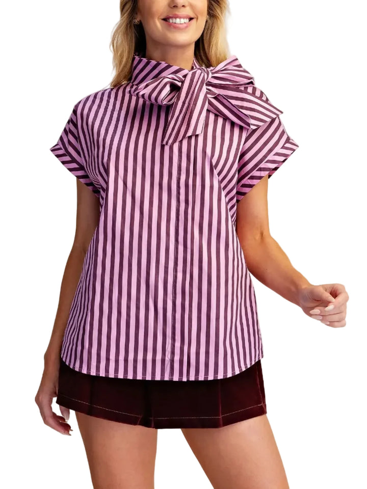 A woman modeling a short-sleeve top with vertical pink and white stripes and a large bow tie neckline, paired with dark shorts, against a plain beige studio background.