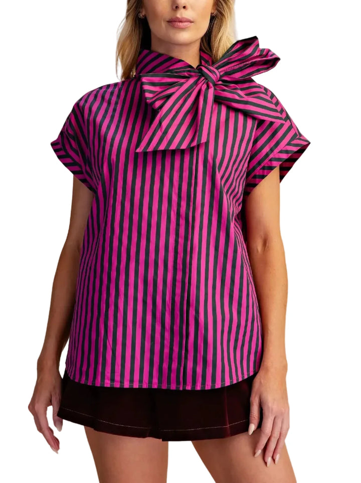 A woman modeling a short-sleeve top with vertical pink and black stripes and a large bow at the collar, paired with dark shorts, against a plain beige studio background.