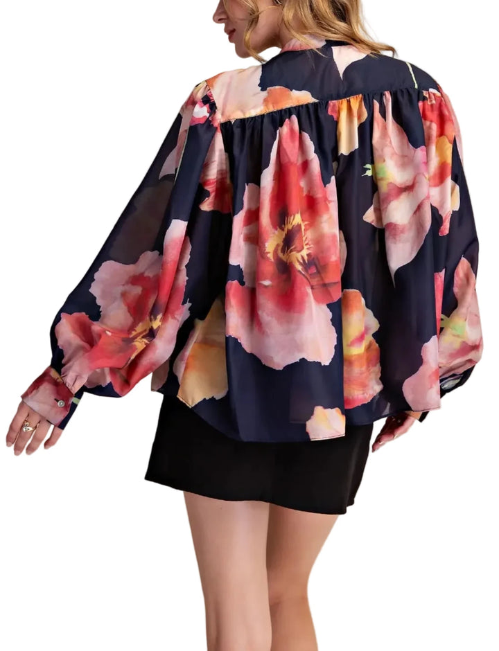 A woman shown from the back wearing a navy blue blouse with a large pink, red, and peach floral print, paired with a black skirt, against a solid beige studio background.