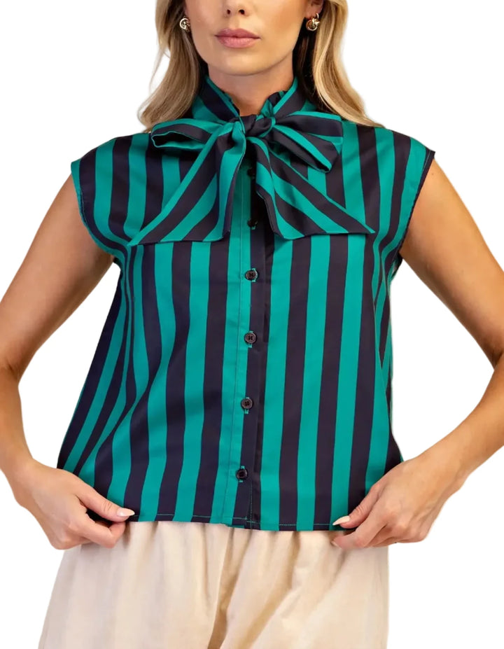 A woman modeling a sleeveless button-front blouse with vertical teal and black stripes and a large bow at the collar, paired with light beige bottoms against a plain beige studio background.