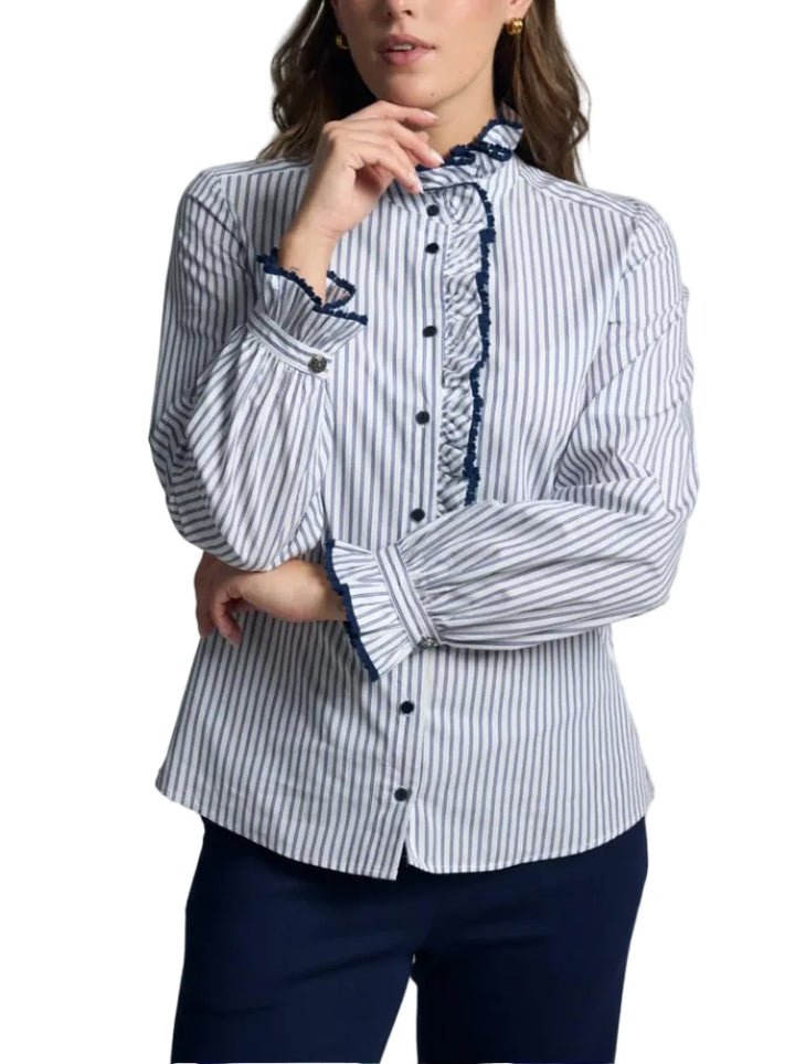 A woman modeling a long-sleeve button-up blouse with vertical navy and white stripes, featuring a ruffled high collar and cuffs, paired with dark navy pants, against a neutral gray studio background.