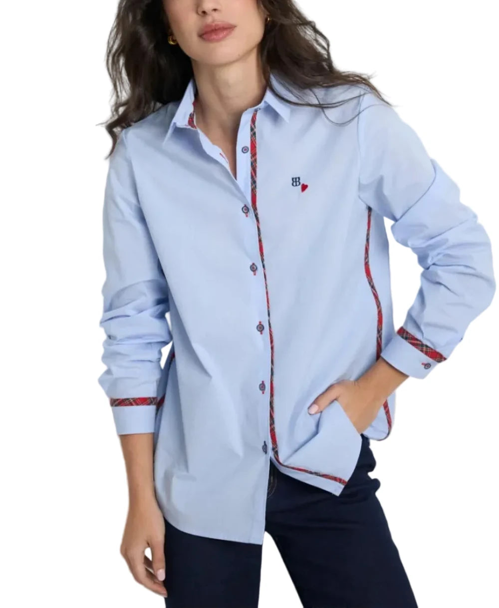 A woman wearing a light blue long-sleeve button-up shirt with red and blue patterned trim details, paired with dark pants, posing against a light gray studio background.