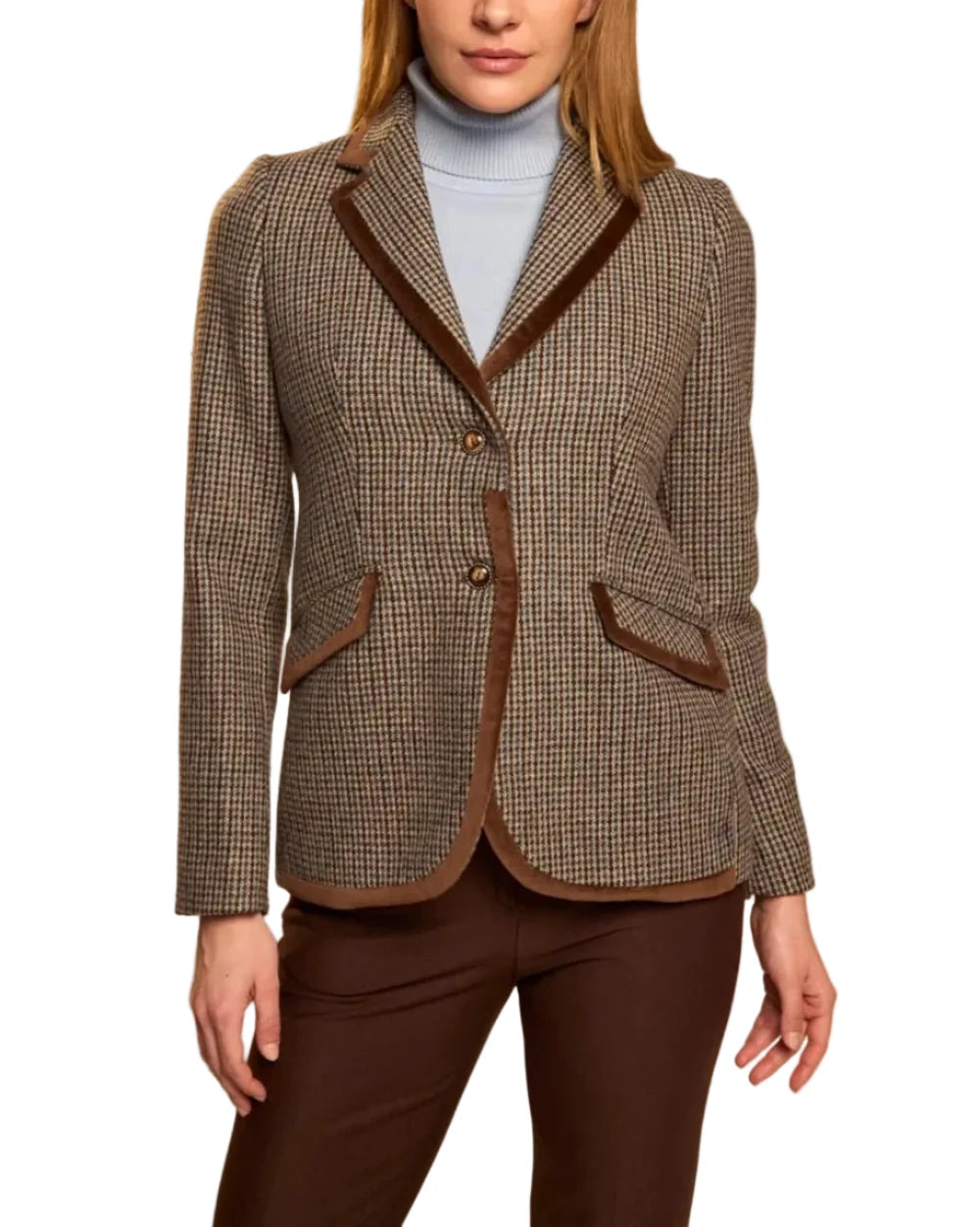 A woman modeling a brown and beige houndstooth blazer with dark brown trim, worn over a light blue turtleneck and dark brown trousers, against a solid beige studio background.