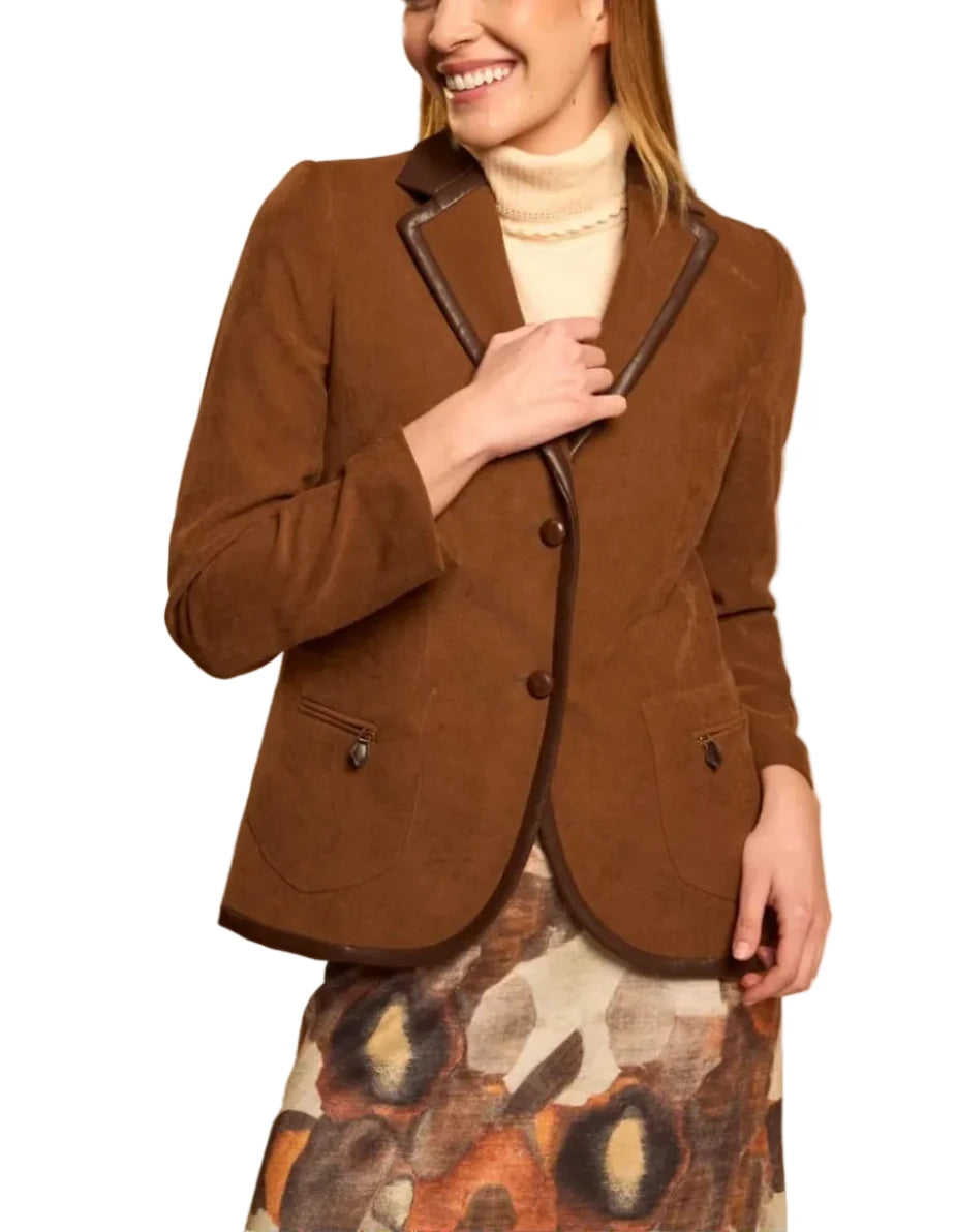 A woman modeling a brown suede-like blazer with dark contrast piping, worn over a cream turtleneck and a patterned skirt, against a solid beige studio background.