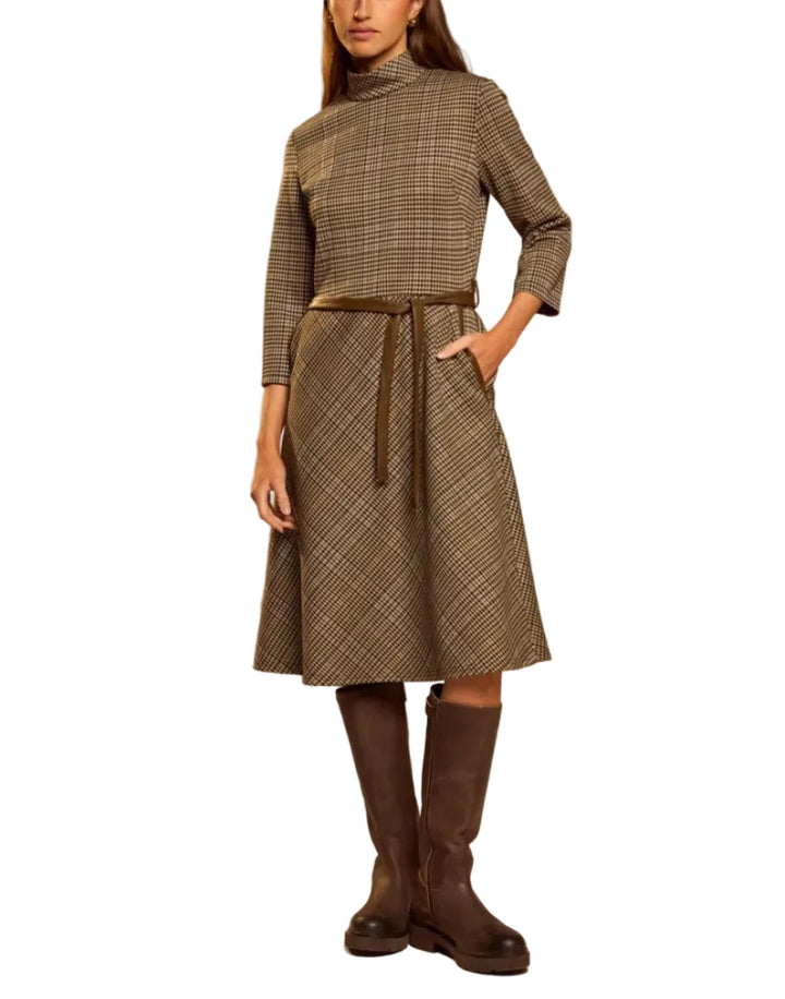 A woman models a mid-length plaid turtleneck dress with three-quarter sleeves and a matching waist tie, styled with dark brown knee-high boots against a solid beige studio background.