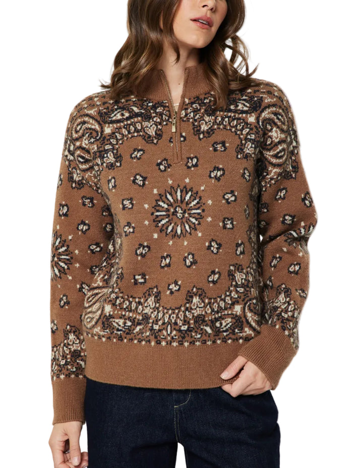A woman wearing a brown knit sweater with a detailed black and beige geometric and floral pattern, paired with dark blue jeans, against a plain light gray background.