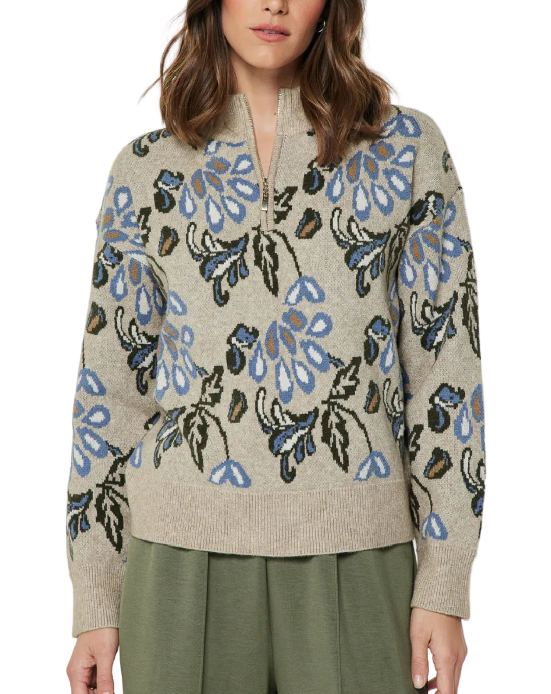 A woman wearing a beige knit half-zip sweater with a blue, green, and black floral pattern, paired with olive green pants, against a plain light gray background.