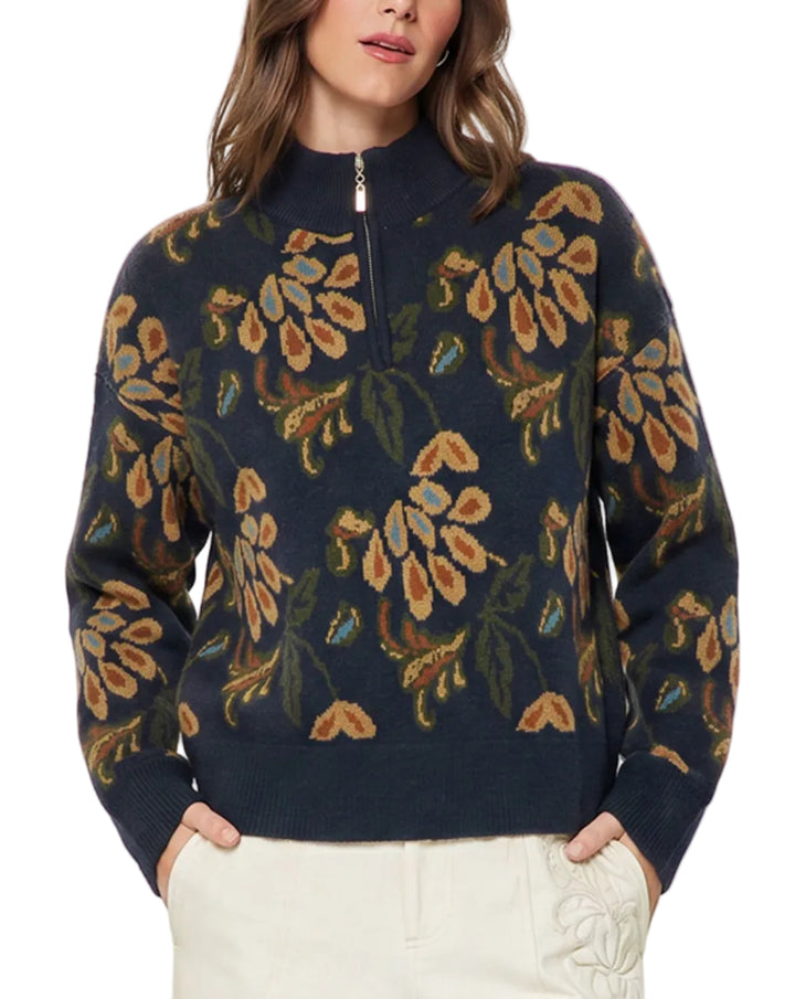 A woman wearing a navy blue quarter-zip sweater with a large floral pattern in tan, olive green, and blue, paired with cream-colored pants, against a plain light gray background.