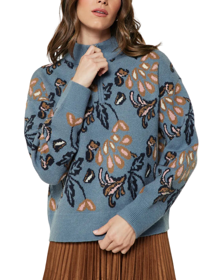 A woman wearing a dusty blue knit sweater with a large floral and leaf pattern in black, brown, and beige, paired with a brown pleated skirt, against a light gray studio background.
