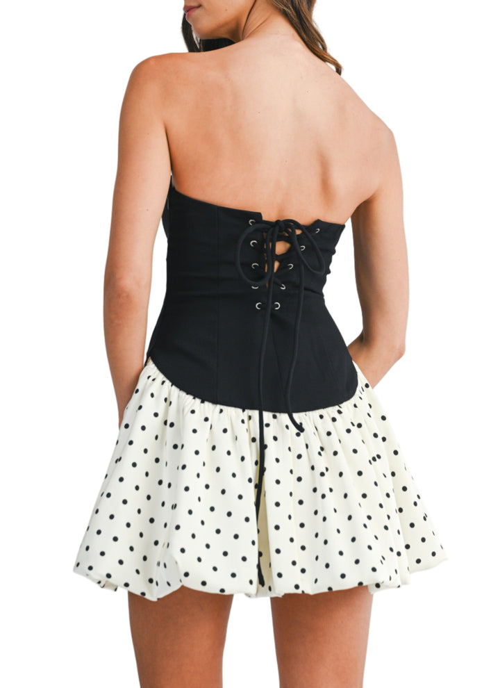 Woman wearing a black strapless top with lace-up back and a white polka dot skirt on a plain background