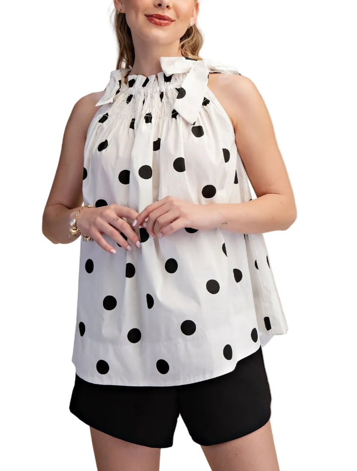 Woman wearing a white sleeveless top with black polka dots and black shorts on a beige background