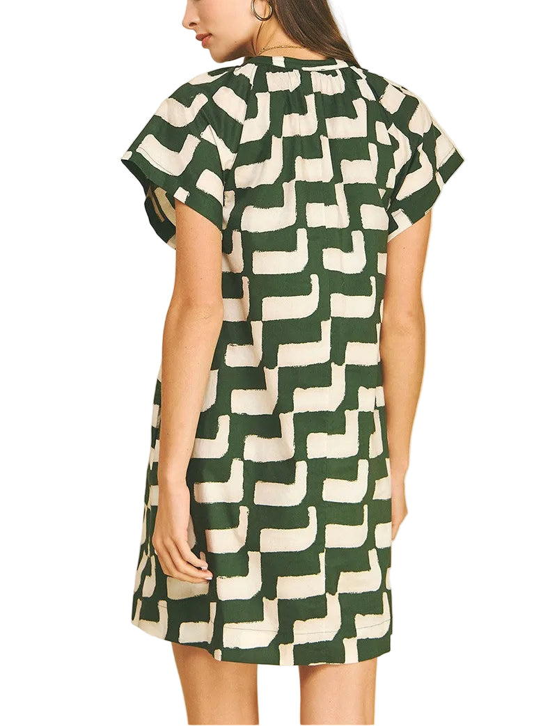 A woman modeling a short-sleeve A-line dress with a dark green and cream geometric block pattern, shown from a rear three-quarter view against a solid beige background.