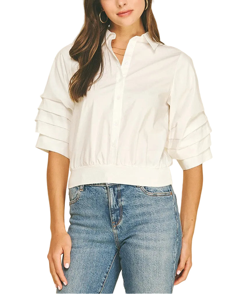 A woman wearing a white short-sleeve button-up blouse with pleated sleeve details and blue jeans, standing against a solid beige background.