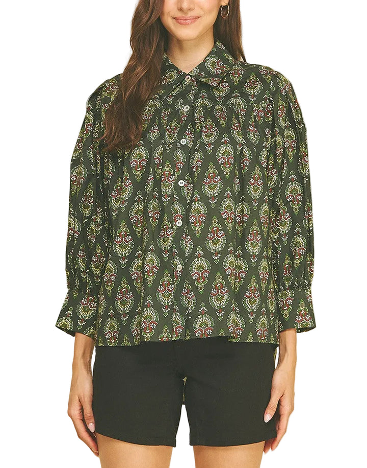 A woman modeling a dark green long-sleeve button-up shirt with a detailed multicolor paisley pattern, paired with black shorts, against a solid beige background.