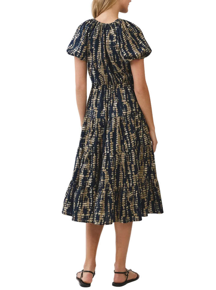 A woman wearing a short puff-sleeve midi dress with a navy blue and beige abstract tie-dye pattern, shown from the back against a light gray studio background.