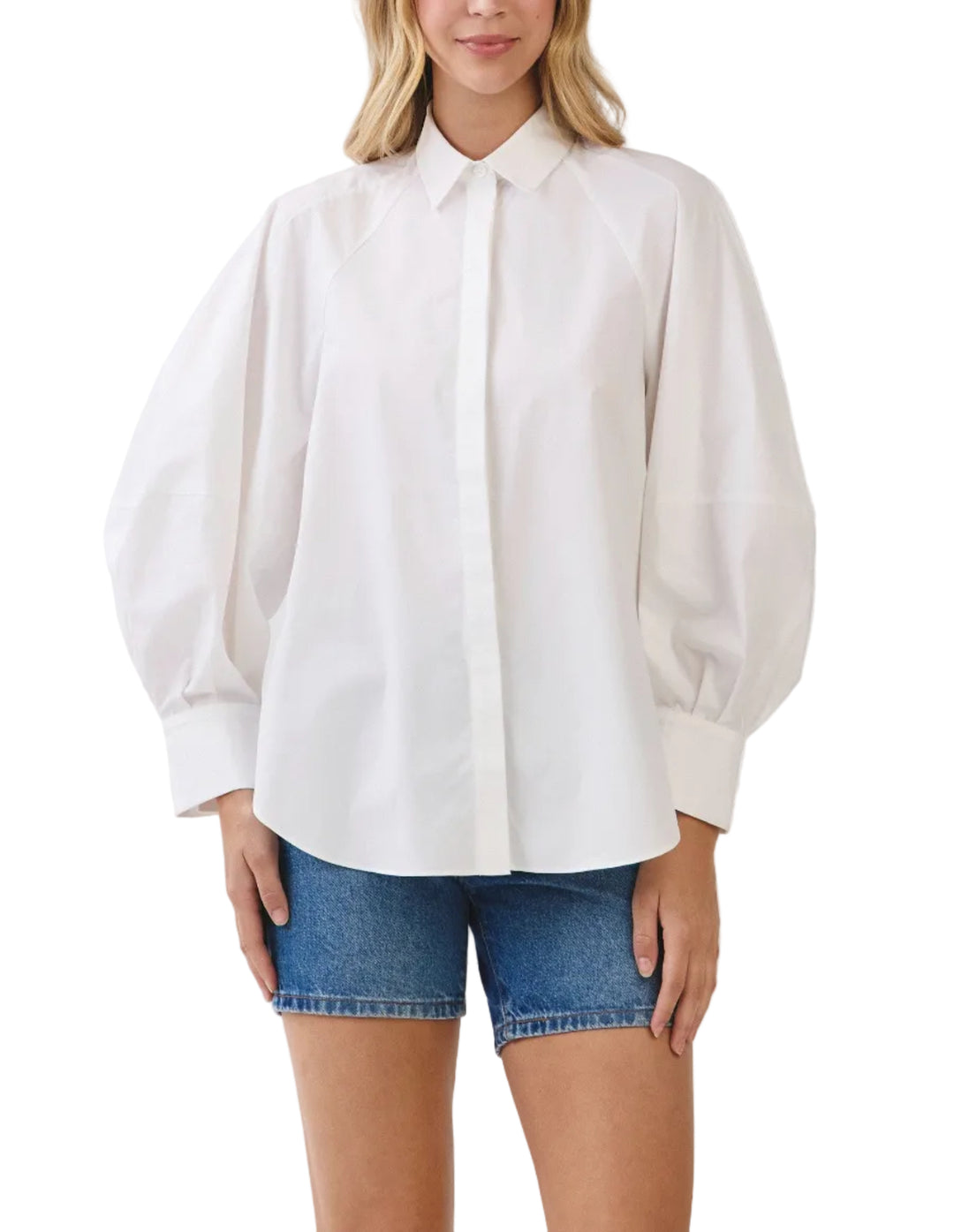 A woman modeling a white long-sleeve button-up blouse with voluminous balloon sleeves and a pointed collar, paired with blue denim shorts, against a light gray studio background.