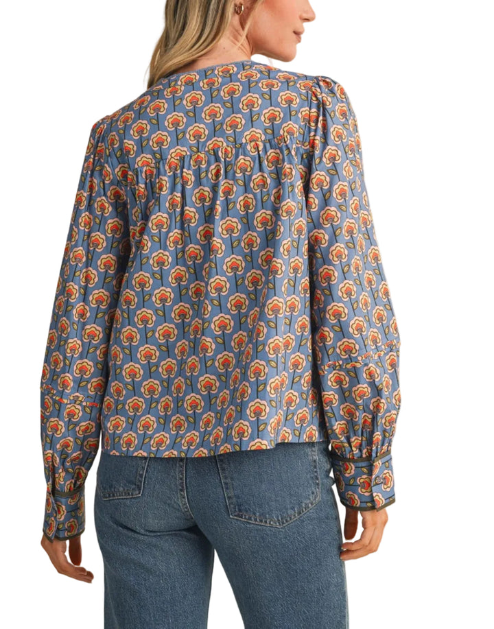 A woman shown from the back wearing a long-sleeve blouse with a blue base and an orange and yellow floral pattern, paired with blue jeans, against a solid beige background.